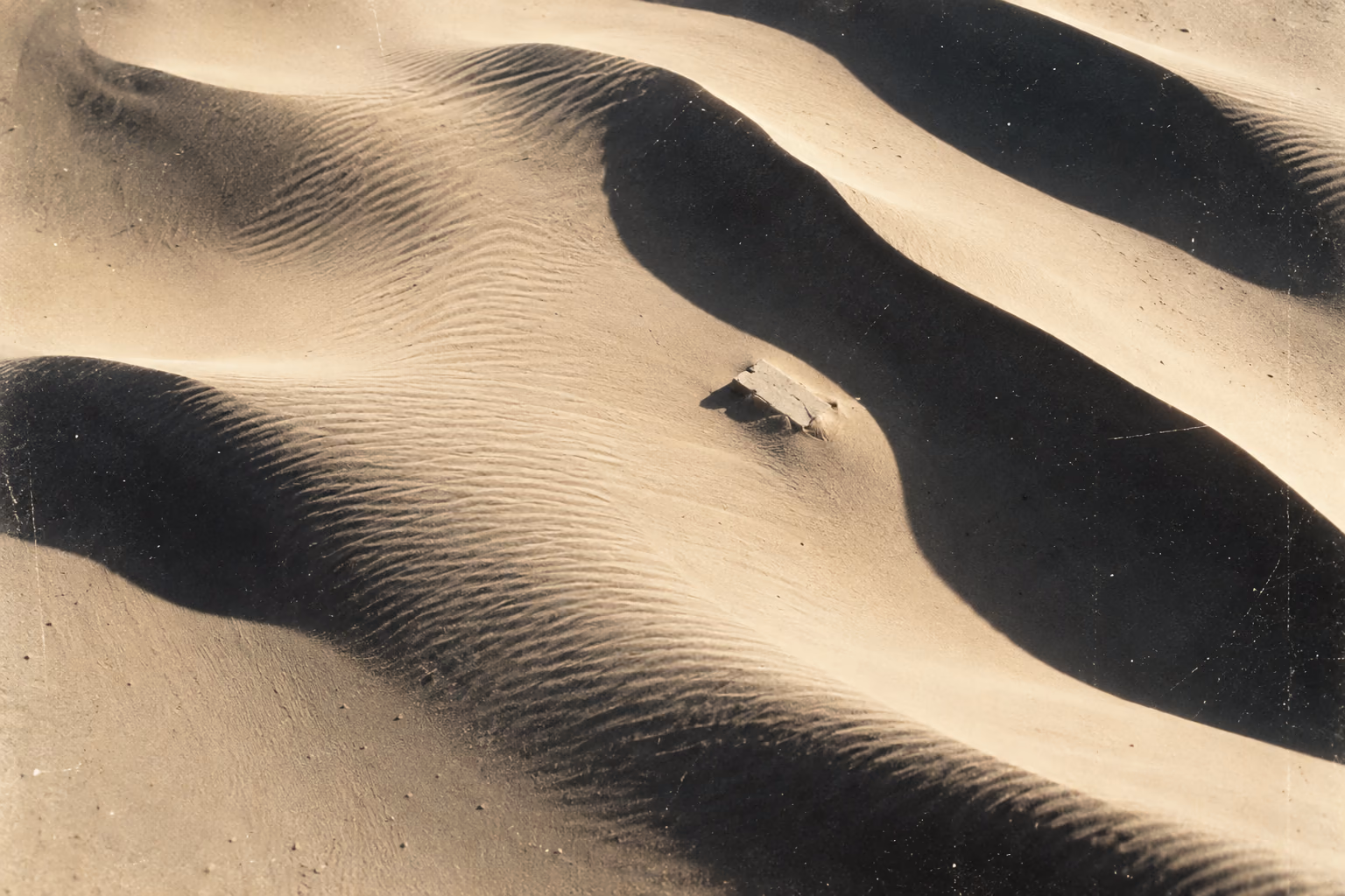 Aerial view of pale dunes with long black shadows and one small concrete slab half buried in the sand.