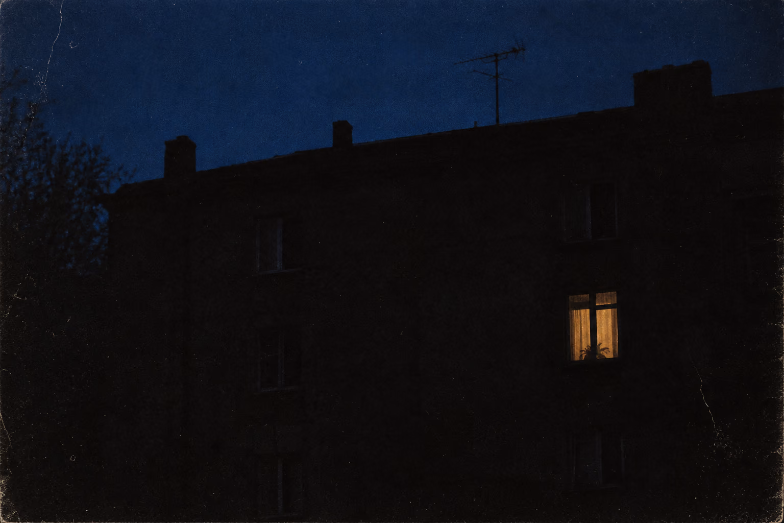 One amber window lit on a dark residential wall at night, a thin antenna against a deep blue sky.